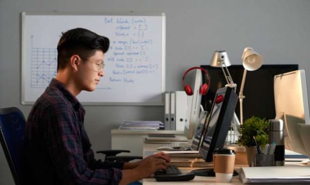 A man with glasses is sitting at a desk looking at a monitor screen with hands resting on the keyboard. The desk has a coffee cup and a full folder.