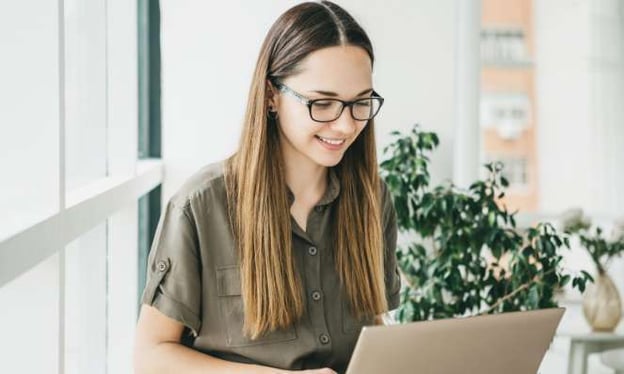 A woman sitting at a desk smiling, working on a laptop. In the background is a plant and a vase. Behind her are windows.