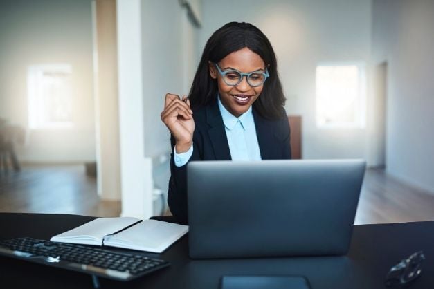 Woman smiling, wearing glasses, seated at desk, looking at laptop with notebook open beside her.