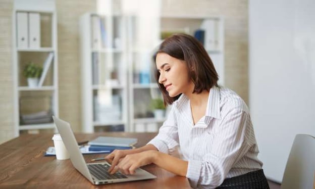 A woman is sitting at a table working on a laptop with a coffee cup and notebooks next to the laptop. She has short brown hair. 