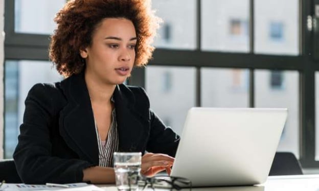 A woman is sitting at a desk working on a laptop. On the table is a glass of water, a notebook, pen, and glasses. The sun is in the window behind her.