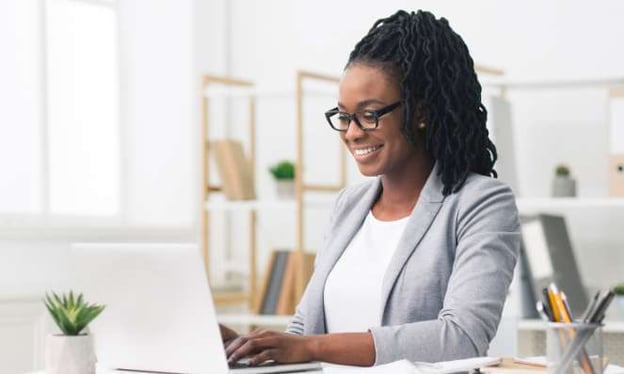 A woman wearing glasses and a gray blazer is sitting at a desk working on a laptop while smiling. On the desk is a small plant and a jar of pens. 