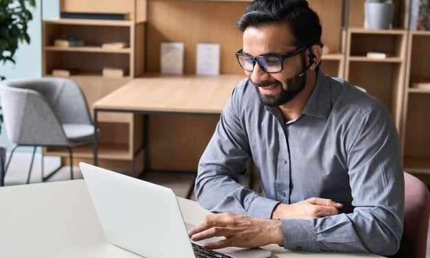 Man seated at desk in office reading laptop smiling
