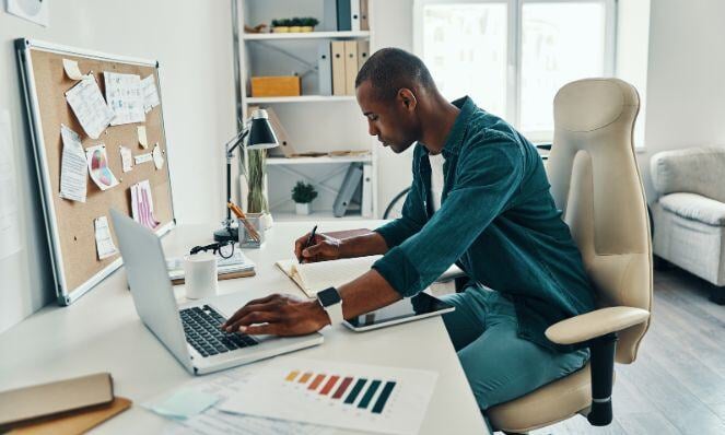 A man sits in a tan-colored chair at a desk with an open laptop. One hand is on the laptop keyboard and the other is writing in a notebook.