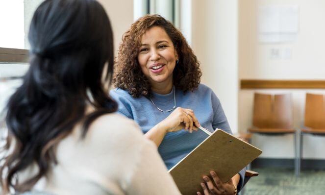 A woman is smiling at the woman she is talking to. They are seated, the smiling woman is holding a pen and clipboard, and the other woman has her back to the camera.