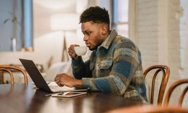 A man is sitting at a table while sipping out of a mug while working on a laptop with a table on the table next to the laptop. 