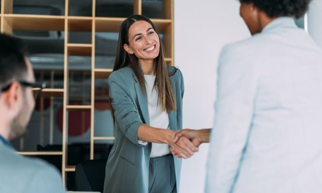 Two women, standing and shaking hands in business attire. A man is seated in the foreground of the image with his back to the camera. He is wearing black eyeglass frames. There are bookshelves in the background of the image.