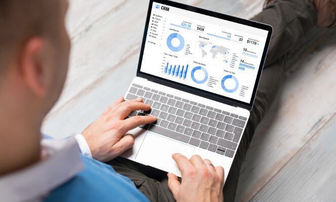 Looking over a person's shoulder as they type on a laptop keyboard. Reports from a CRM are visible on the laptop screen.