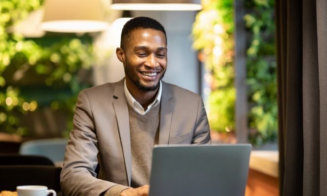 An African American man is seated at a table with a coffee cup and a laptop on it. He is smiling at the laptop. There are biophilic design walls in the background of his office.