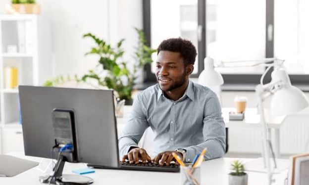 A man sits at a desk while smiling and looking at a computer monitor with his hands typing on the keyboard. There are plants and windows behind him.