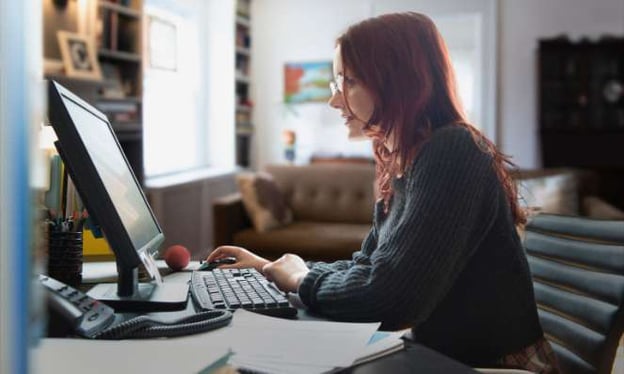 A woman is sitting at a desk looking at a computer screen. The desk has a phone, a ball, and stacks of paper on it.