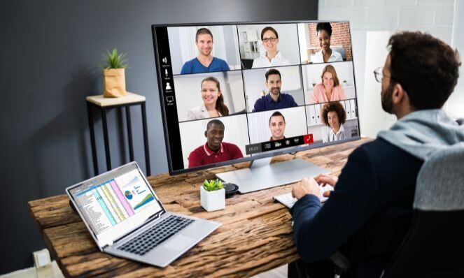 A man wearing glasses sits at a desk with a laptop (showing reports) and a monitor with many faces on a Zoom call.
