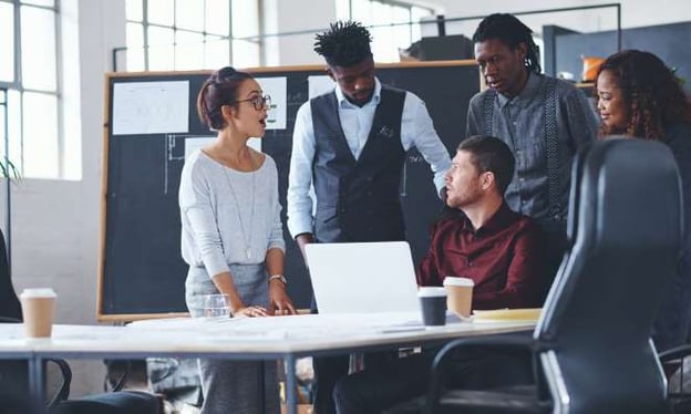 A group of 5 people. 4 are standing and 1 is sitting at the table. All members are in a discussion. There is an open laptop on the table. 