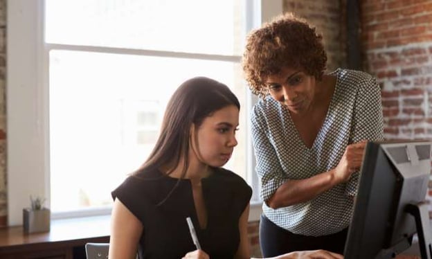 A woman is sitting at a desk looking at a monitor while another woman is standing next to her and looking at the monitor screen. 