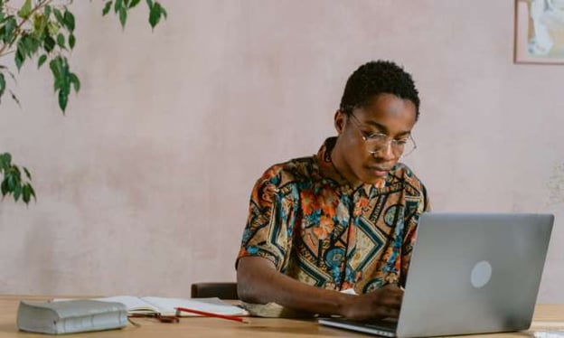 A man is wearing glasses and sitting at a table while working on his laptop. On the table is a pen, book, and notebook.