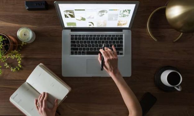 An overhead view of a table with two female arms. One arm is on an agenda open on the table, and the other is resting on an open laptop.