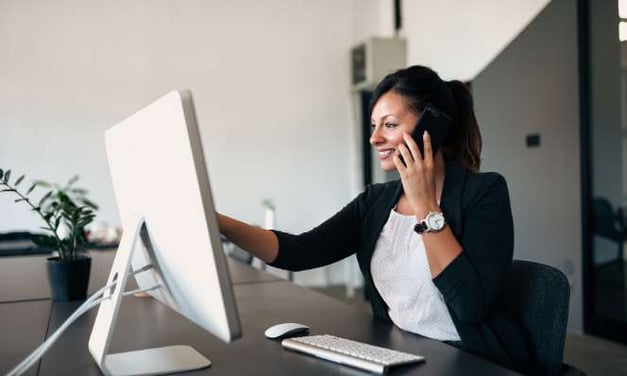 A woman sits at a desk, talking on her cell phone while looking at her computer monitor. She has dark hair and is wearing a black blazer and a watch. 