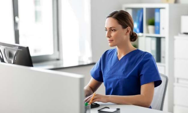 Woman sitting at a desk, looking at a computer monitor, typing on a keyboard. She has her hair in a bun and is wearing a blue shirt. 