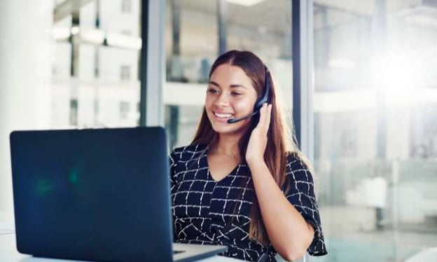 A woman sitting at a desk smiling while looking at a laptop screen with a headset on. She is wearing a black top with white lines. 