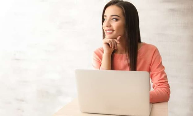 A woman is sitting at a desk smiling, looking to her right with a laptop on the table in front of her and her head resting on her hand. 