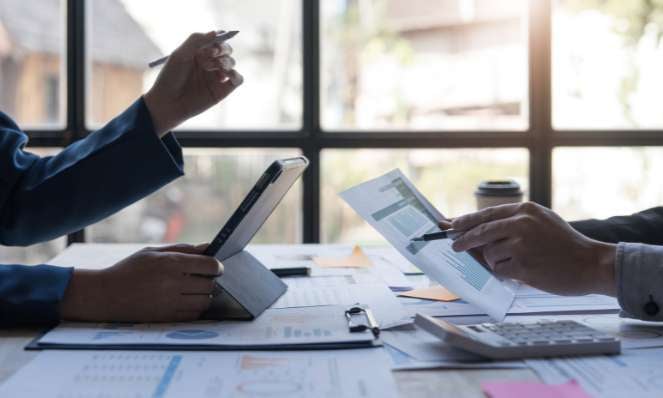 Two people at a desk with papers and a tablet. They are both holding pens. There is a window behind them. Only their hands and arms are visible.