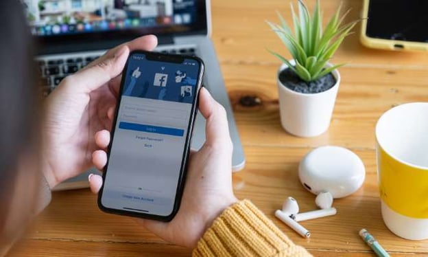 Hands holding a cell phone with the Facebook login screen pulled up are seated at a desk with a laptop, air pods, a pencil, a coffee cup, and a plant.