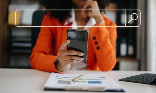 A woman is using her phone to do a voice search while at work. There is a clipboard with reports on it along with a pen on the desk in front of her.