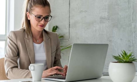 A woman with glasses and a coffee cup is working at a laptop on a desk. There is a potted plant on the desk in the foreground, a window in the back. 