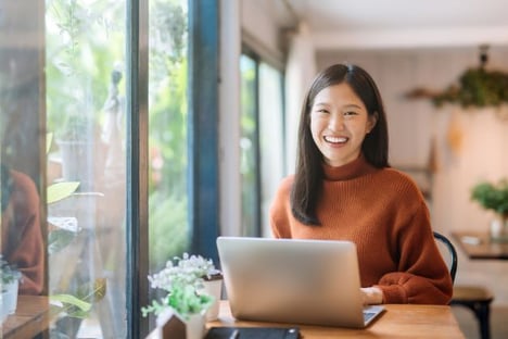 Woman seated at a table, using laptop, smiling, windows next to her.