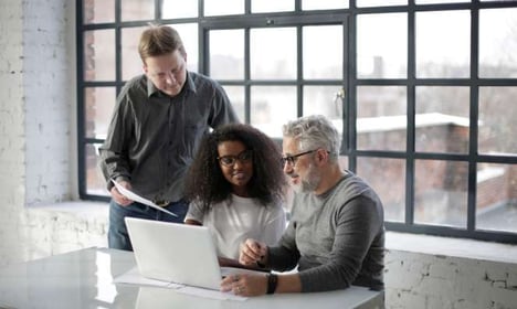 Three people looking at a laptop screen, two seated, one standing with papers in his hand. Windows are behind them in the background.