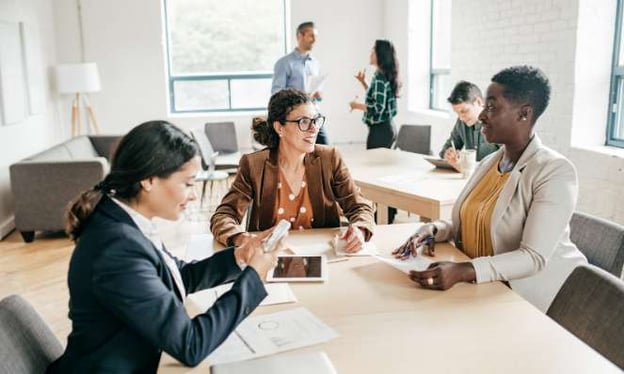 Three women seated at a table, there is a tablet on the table. Three other people are in the background, one reading alone, two standing speaking.