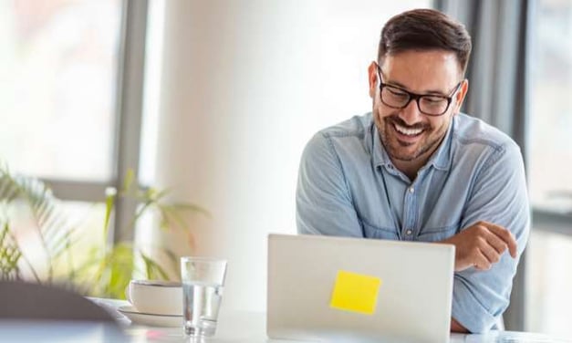 Man smiling at a desk, looking at a laptop screen. The desk also has a glass of water on it. The laptop has a sticky note on the back of the monitor.