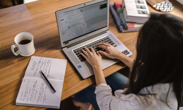 A woman is sitting at a table with her hands on a laptop keyboard and the screen facing the camera. On the table is a cup of coffee and a notebook.