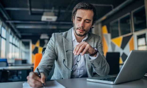A  man in a grey suit is sitting at a desk working. He is writing on a notepad with one hand and holding glasses with the other. 