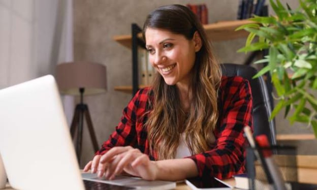 A woman wearing a red plaid sweater is sitting at a desk working on a laptop. On the desk are also a phone and a jar of pens. 