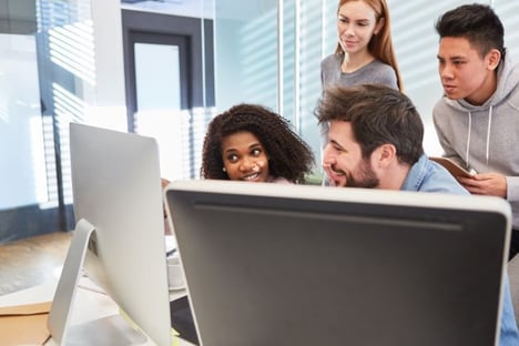 Four people looking at computer monitors, two seated, two standing.