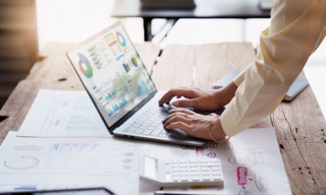 A person is standing over a laptop which is placed on a wooden table. The laptop has reports on the screen. There are papers and a calculator also on the table. The person is typing.