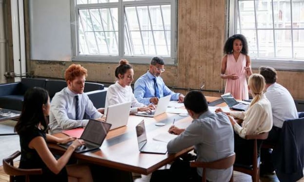 Seven people seated around a board room table, one woman standing speaking to the group. Many laptops open on the table. Windows in background.