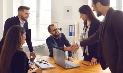 Five people in a room, two seated at desk, three standing, wearing business attire. A laptop is open on the desk along with a notepad with glasses.