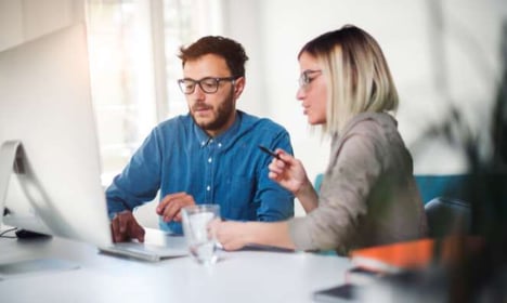 Two people seated at a desk, looking at a computer monitor. 