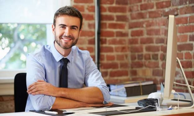 A man with crossed arms is working at a desk, smiling while working on a desktop. He is wearing a light blue dress shirt and dark tie.