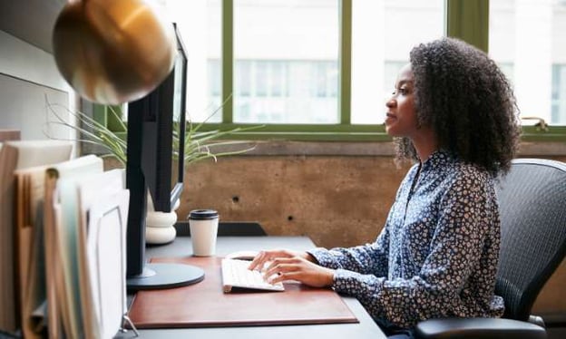 A woman sits at a desk looking at a monitor while typing on a keyboard. A lamp, a stack of folders, and a disposable coffee cup are on the desk.