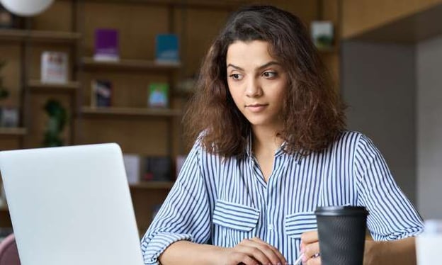 A woman in a blue and white striped shirt is sitting at a desk working on a laptop. She is writing a note and has a coffee cup next to her. 