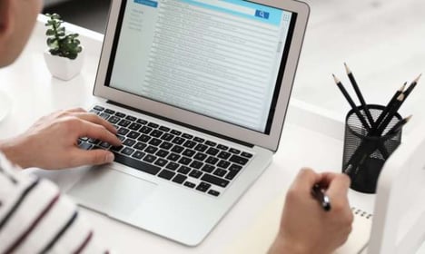 Person seated at desk, one hand on laptop, opening emails, the other holding a pen to write on a notepad.