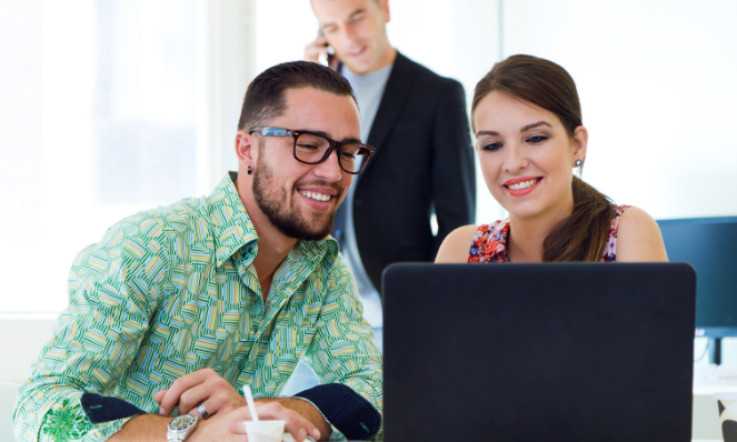 Two people are seated, smiling, and looking at a laptop screen. A man in the background is standing and talking on his cell phone.