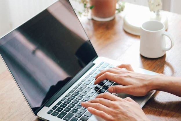 Hands typing on a laptop which is placed on a wooden table. A coffee mug is to the right of the laptop, and there is a potted plant.