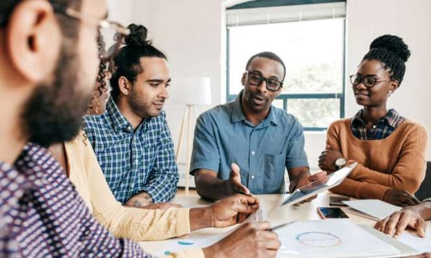 Six people seated around a table, reviewing statistics on paperwork and a tablet the man in the center is holding. There is a window behind him.