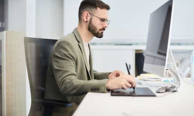 A man wearing glasses is seated at a desk and looking at a computer monitor. He has one hand on a pen and tablet.