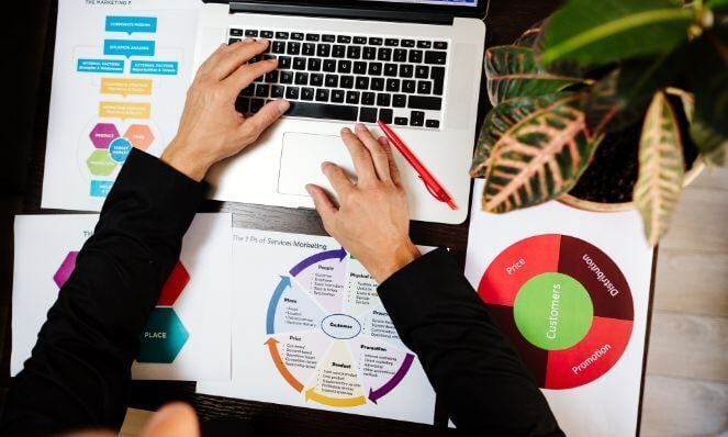 Person typing on their laptop with papers with graphs and data scattered on their desk.