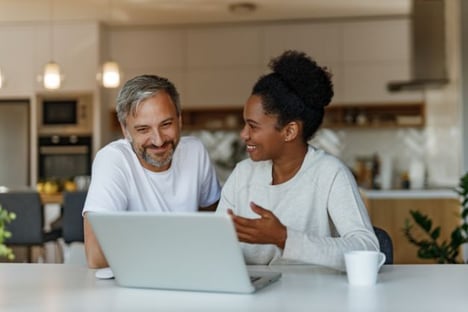 Two people seated at a table, one looking at the other, the other looking at a laptop screen. Coffee cup in the foreground, a kitchen in background.